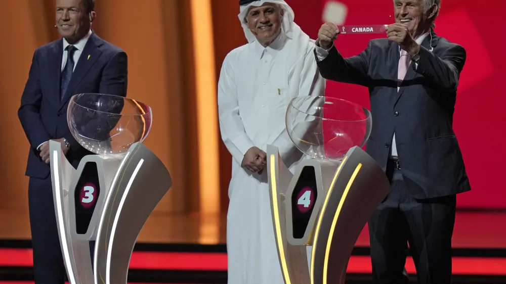 Former German soccer international and manager Lothar Matthaus, left, and former Qatari soccer international Adel Ahmed MalAllah look on as Serbian-Mexican retired soccer manager Bora Milutinovic holds up the name of Canada as he assists in the 2022 soccer World Cup draw at the Doha Exhibition and Convention Center in Doha, Qatar, Friday, April 1, 2022. (AP Photo/Darko Bandic)