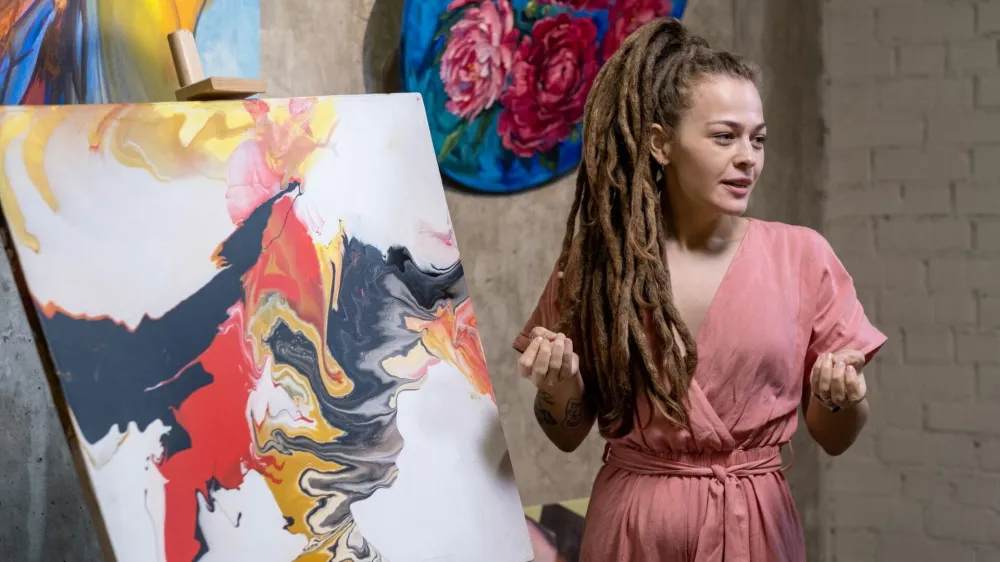 Young female auctioneer or speaker with dreadlocks describing abstraction illustrated on painting in front of audience