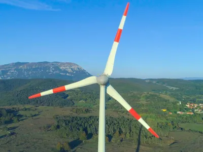 AERIAL CLOSE UP: Spring sunrays illuminating white metal blades in green countryside. Wind-turbine generator spinning in strong winds, representing a renewable source of electricity for village nearby