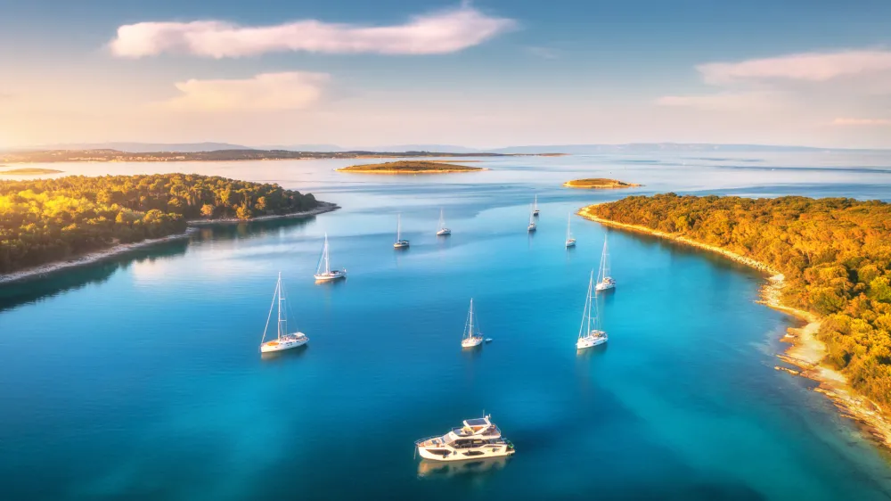 Aerial view of beautiful yachts and boats on the sea at sunset in summer. Adriatic sea, Kamenjak, Croatia. Top view of luxury yachts, sailboats, lagoon, clear blue water, and green forest. Travel