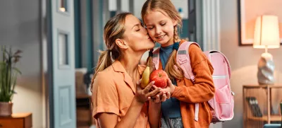 Healthy snack for pupil. Adult woman giving pretty daughter with pink schoolbag apple and banana and kissing her in cheek. Casual female taking care about nutritious meal between lesson for child. / Foto: Harbucks