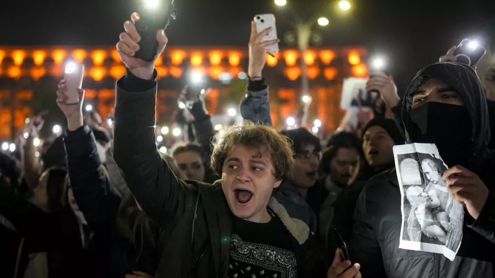Youngsters shout slogans and flash the light of their mobile phones in Bucharest, Romania, Wednesday, Nov. 27, 2024, next to an altered version of a classic painting, depicting Russian President Vladimir Putin and Calin Georgescu, the independent candidate for Romanian presidency who won the first round of elections making it to the Dec. 8, runoff. (AP Photo/Vadim Ghirda)
