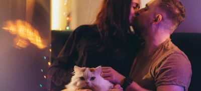 A couple of a guy and a girl are sitting on a bed in a room with colored lighting. A guy and a girl kiss. A girl has a white cat sitting in her arms / Foto: Illiak