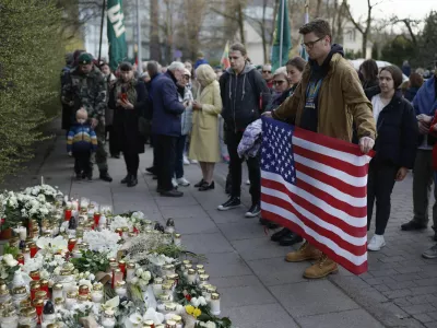 A man with a U.S. national flag and other people stand as they come to pay their last respects in memory of three U.S. Army soldiers found dead on Monday at a training range in Pabrade, near the U.S. Embassy in Vilnius, Lithuania, Tuesday, April 1, 2025. (AP Photo/Mindaugas Kulbis)
