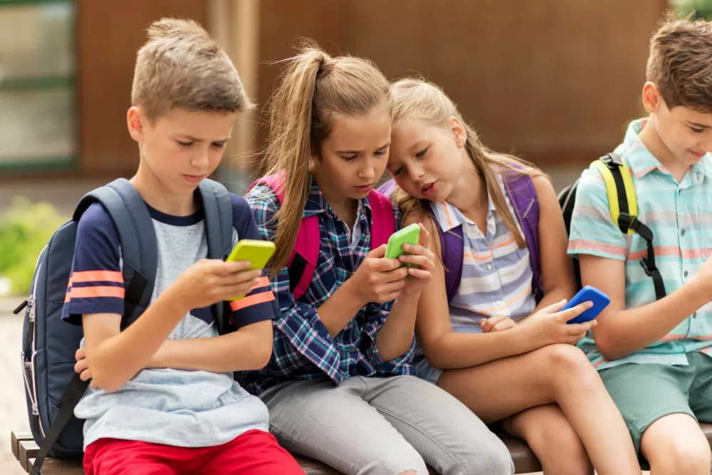 primary education, friendship, childhood, technology and people concept - group of happy elementary school students with smartphones and backpacks sitting on bench outdoors / Foto: Dolgachov