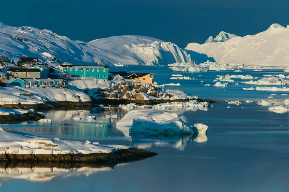 Colorful houses line the shores of a serene bay in the Arctic, surrounded by icebergs and snow-covered mountains, reflecting the warm glow of the sunset on the water. / Foto: Mikael Svensson