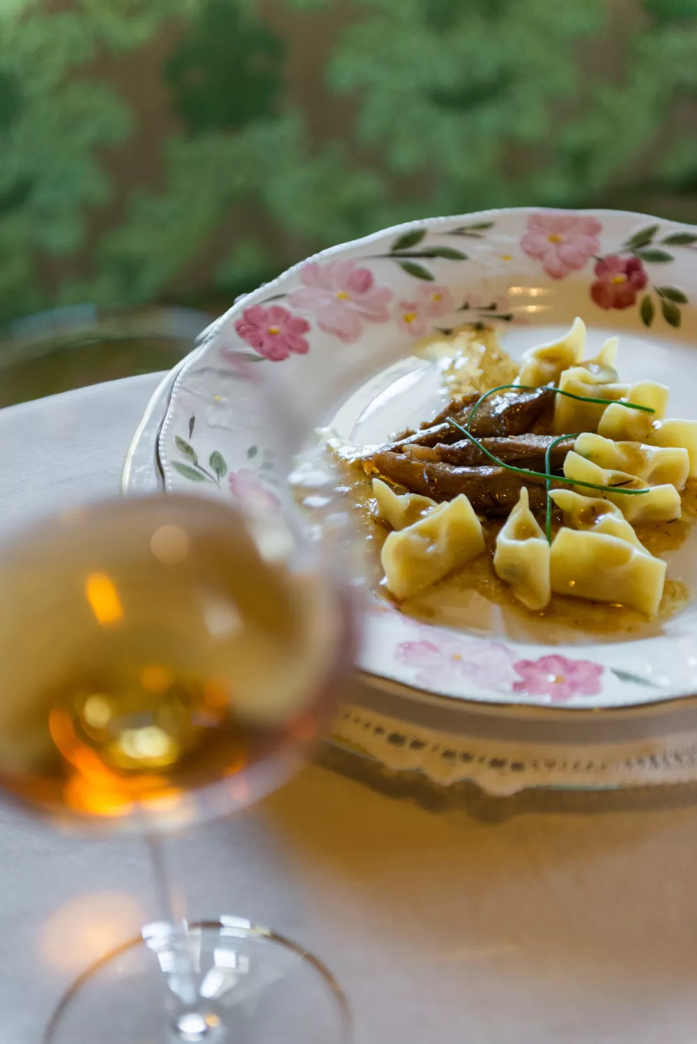 A composition of a traditional gourmet dish (from Idrija, Slovenia), žlikrofi with beef, served on a plate (decorated with a floral print), with a glass of wine on the side. The pasta is filled and can be compared to ravioli. The composition is presented indoors on a table. Vertical color images with copy space.