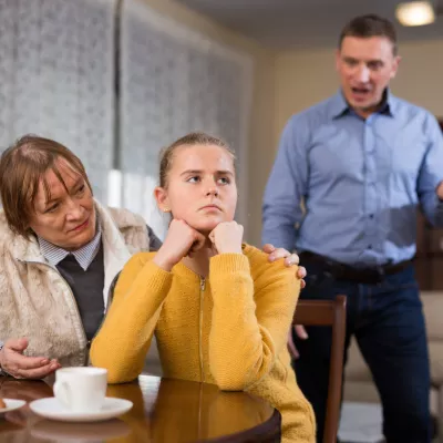 Portrait of troubled teen girl sitting at home table, scolded by her father and grandma / Foto: Jackf
