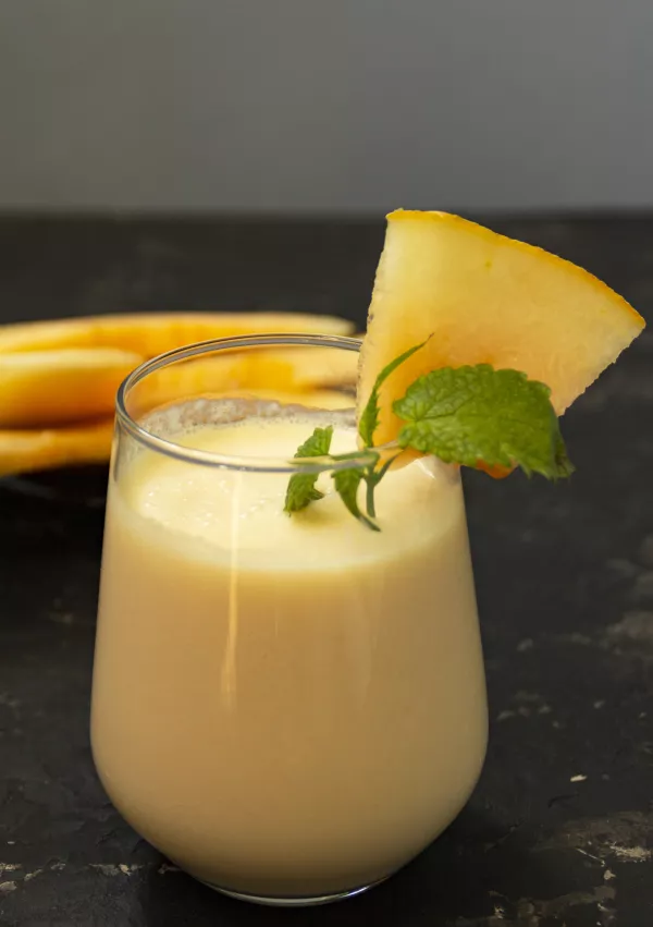 Melon milk shake on a black background. Ripe sliced melon is lying nearby. The smoothie glass is decorated with a sprig of mint and melon crumbs. Summer healthy food concept. / Foto: Venera Chernyshova