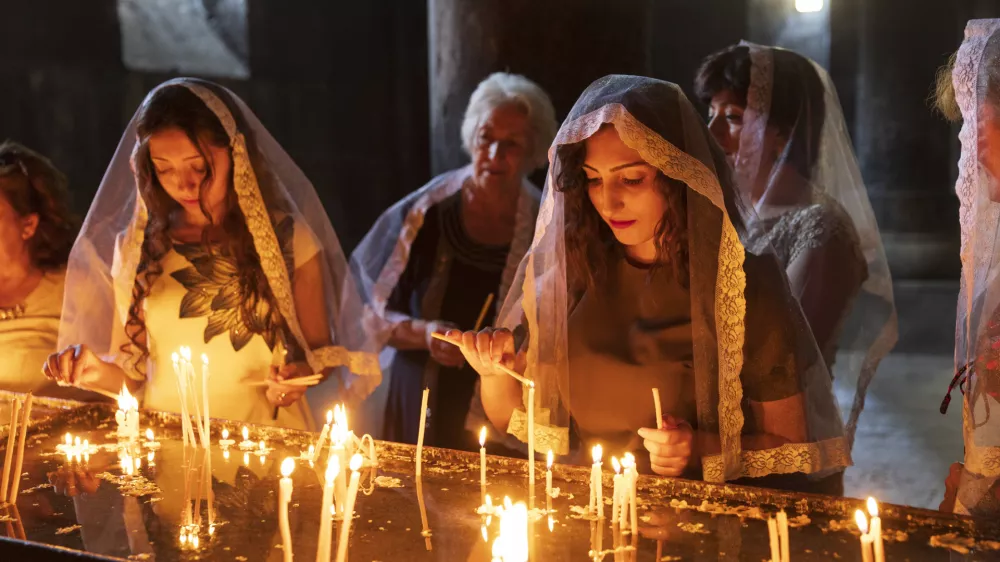August 18, 2018. Armenia, Ararat Province. Geghard Monastery interior. Young women in headscarfs or shawls lighting candles.