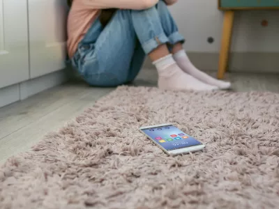 Mobile phone lying on the floor with unrecognizable sad little girl sitting behind. Selective focus on mobile in foreground