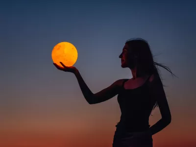 A young woman holds the full moon in her hands against the backdrop of a red sunset. Millennial blogger leads social media. Girl silhouette portrait. Astrology, Waxing Crescent. Copy space / Foto: Julia Pavaliuk