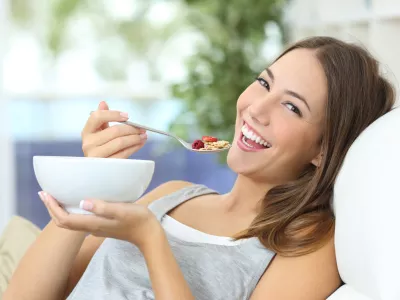 Happy girl eating cereals with fruits from a bowl sitting on a couch at home