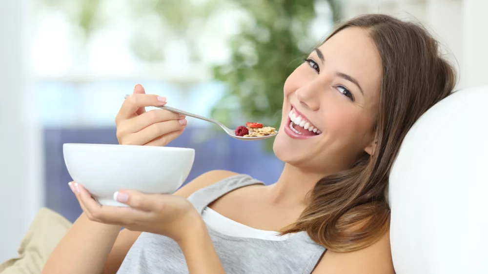 Happy girl eating cereals with fruits from a bowl sitting on a couch at home