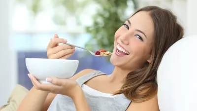 Happy girl eating cereals with fruits from a bowl sitting on a couch at home