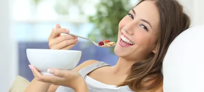 Happy girl eating cereals with fruits from a bowl sitting on a couch at home
