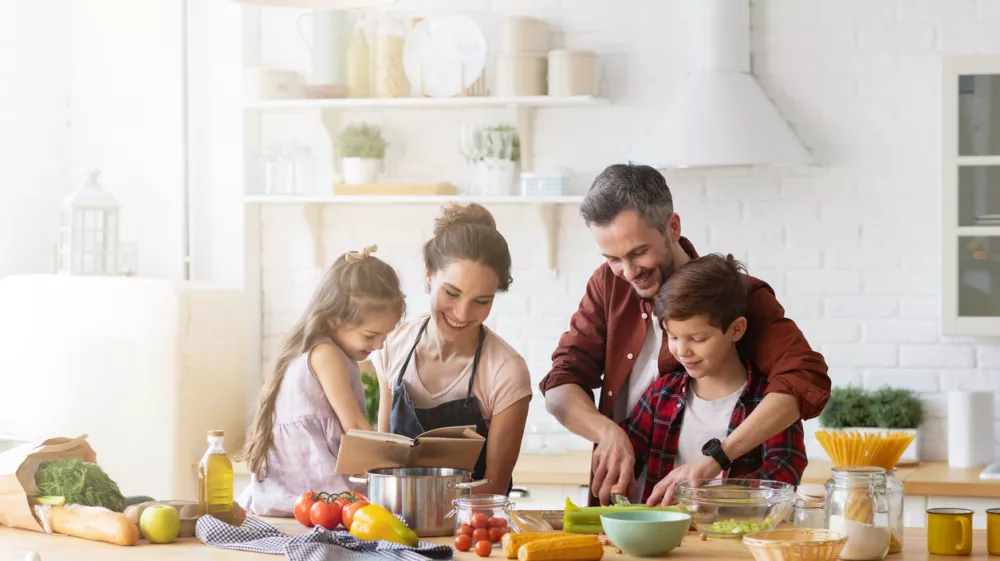 Happy family cooking together on kitchen. Mother and daughter reading recipe to father and son. Dad and boy chopping green vegetable leaf for salad. Home recreation and food preparation on weekend