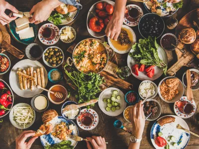 Turkish breakfast table. Flat-lay of peoples hands taking pastries, vegetables, greens, olives, cheeses, fried eggs, spices, jams, honey, tea in copper pot and tulip glasses, wide composition