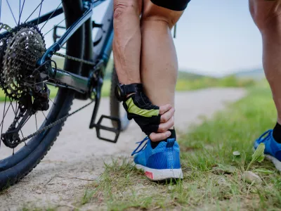 A close-up of active senior man in sportswear suffiering from pain in his ankle after cycling, in park in summer.
