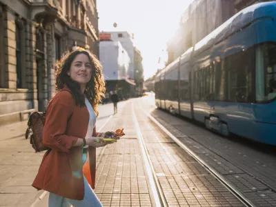 Woman with phone and sunglasses walking across city street next to blue tram. Concept: urban lifestyle, travel experience, exploring Croatia.