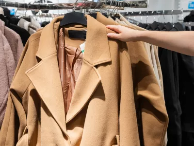 A shopper's hand selecting a stylish camel coat from a rack filled with various garments in a retail clothing store / Foto: Volha Van Meer