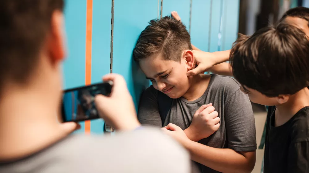 A young boy is being bullied by peers at school. The boy is upset as another boy grabs his neck. A third boy records the bullying incident on his phone. Bullying of teenager in high scool.