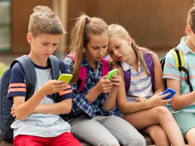 primary education, friendship, childhood, technology and people concept - group of happy elementary school students with smartphones and backpacks sitting on bench outdoors