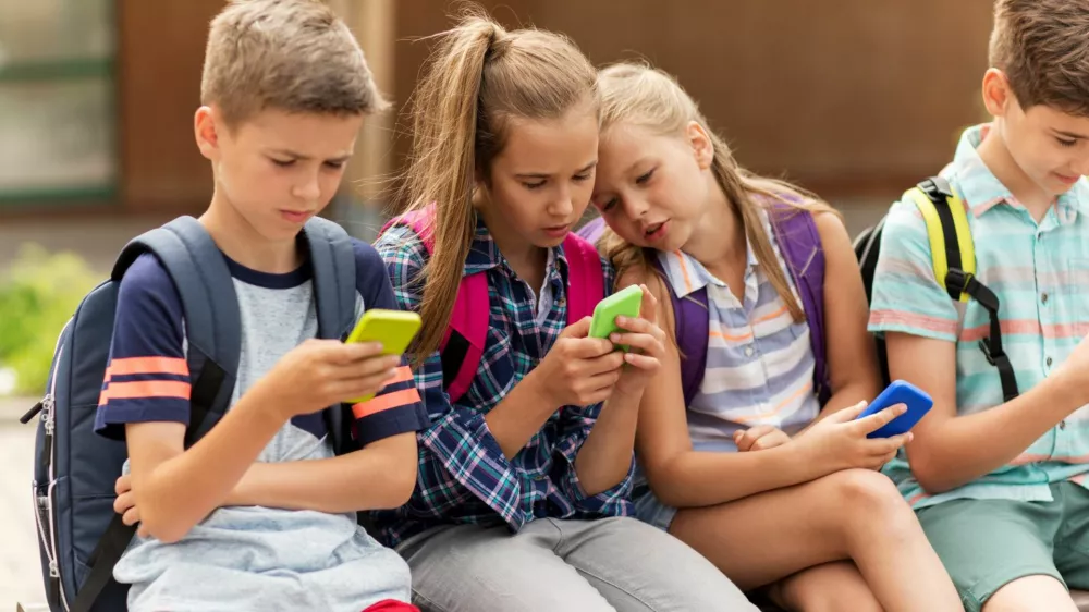 primary education, friendship, childhood, technology and people concept - group of happy elementary school students with smartphones and backpacks sitting on bench outdoors