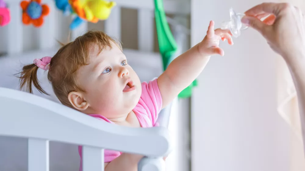 Little girl pulls her hand to the dummy, standing in a baby crib