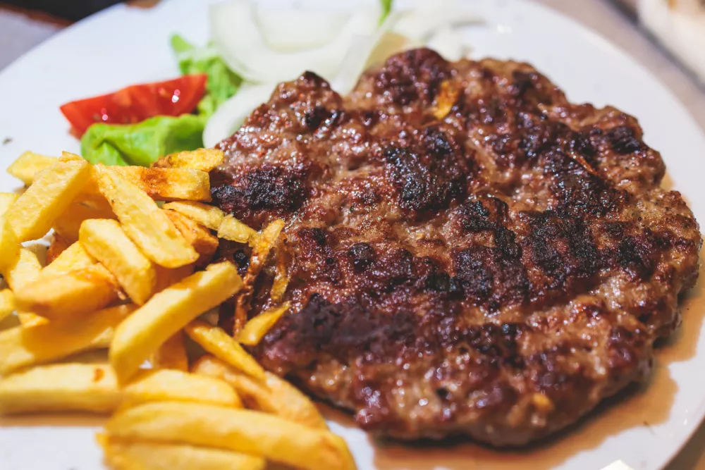 Pljeskavica with french fries on a plate, traditional serbian dish, grilled dish of spiced meat patty mixture of pork, beef and lamb, popular balkan steet food Pleskavitza, served in a cafe / Foto: Nikolay Tsuguliev