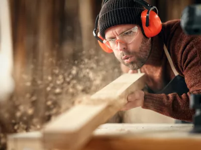 Adult male woodworker in protective goggles and headphones blowing sawdust from wooden detail while working in carpentry workshop / Foto: Evgenyatamanenko