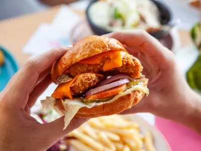 Hands holding fried chicken burger in brioche potato bun in queso sauce.