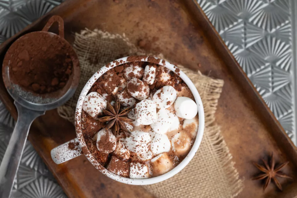 White mug of brown hot chocolate with marshmallows, cocoa powder, and star anise on burlap and a stoneware tray.