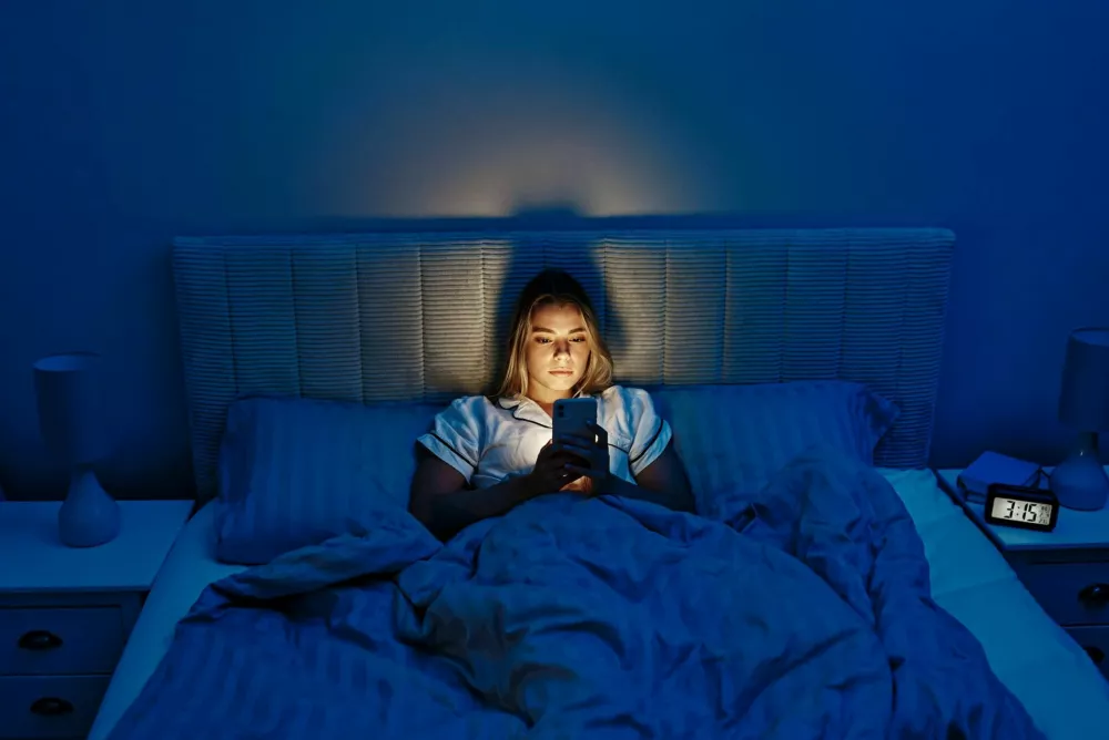 As night falls, a young woman finds solace in her cozy bed, softly illuminated by her smartphone, reflecting the essence of modern life and digital connections that define todays world / Foto: Valerii Apetroaiei
