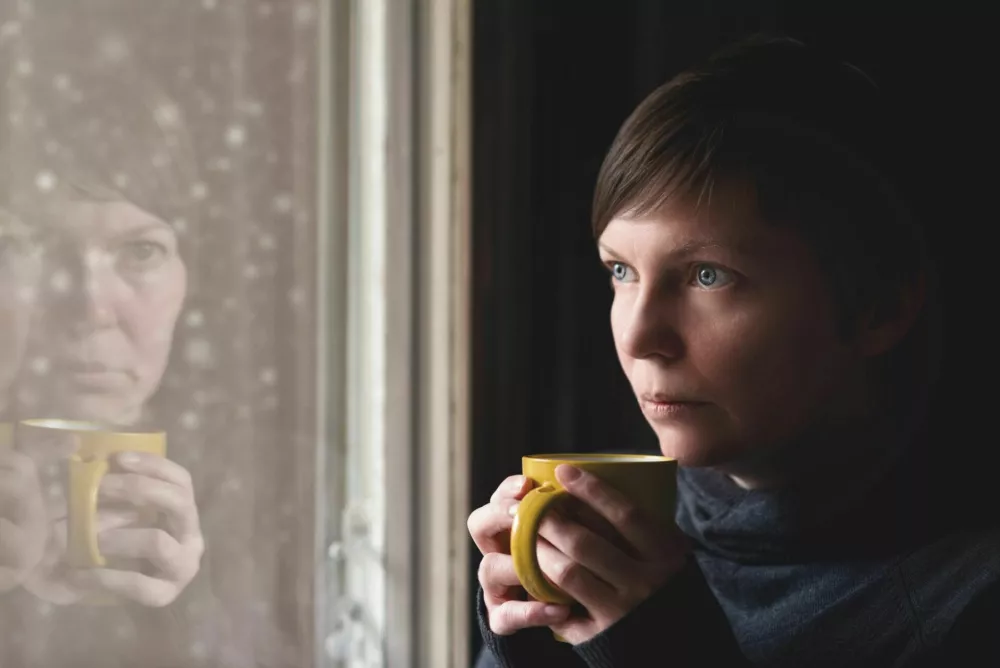 Lonelsome woman drinking cup of coffee by the window of her living room, looking out at snow falling with a sad look on her face. Selective focus with shallow depth of field. / Foto: Stevanovicigor