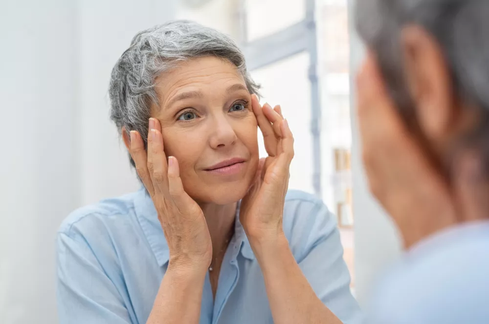Beautiful senior woman checking her face skin and looking for blemishes. Portrait of mature woman massaging her face while checking wrinkled eyes in the mirror. Wrinkled lady with grey hair checking wrinkles around eyes, aging process. / Foto: Ridofranz