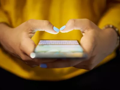Young woman using cell phone to send text message on social network at night. Closeup of hands with computer laptop in background / Foto: Diego_cervo,getty Images/istockphoto