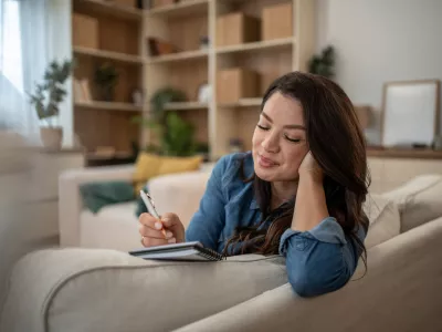 Young woman feeling happy and relaxed while writing personal notes in a diary or journal, reflecting on her day in the comfort of her cozy home environment / Foto: Mirjana Pusicic, Getty Images