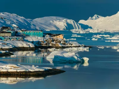 Colorful houses line the shores of a serene bay in the Arctic, surrounded by icebergs and snow-covered mountains, reflecting the warm glow of the sunset on the water.