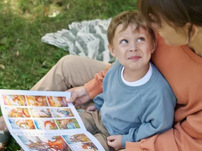 High angle closeup of smiling young boy sitting on mothers lap and looking up playfully while reading book outdoors in green park copy space