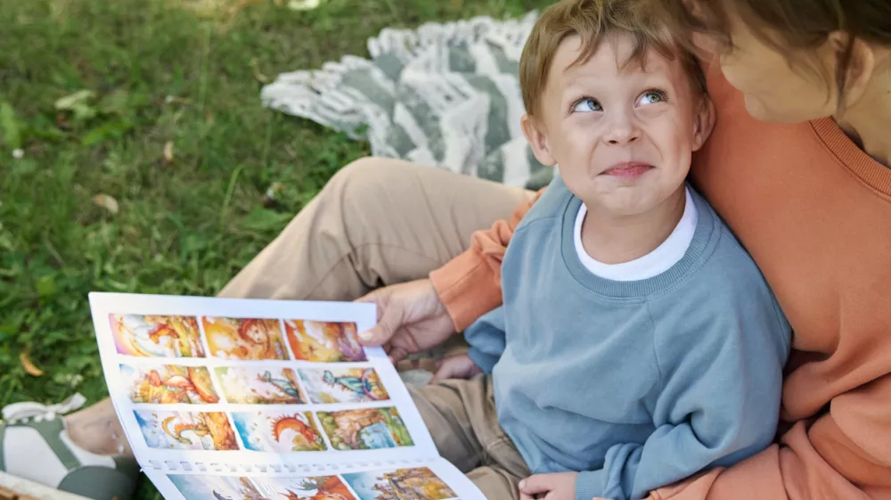 High angle closeup of smiling young boy sitting on mothers lap and looking up playfully while reading book outdoors in green park copy space