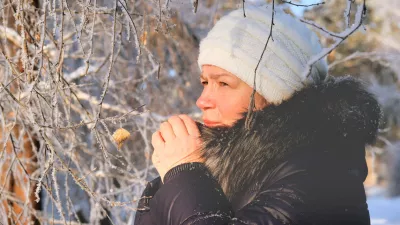 Woman in the blue padded jacket and white cap under snowfall at the street. Pensive attractive mature woman with a quiet smile. Sunny winter vacation outdoors. Deep breathing freshness