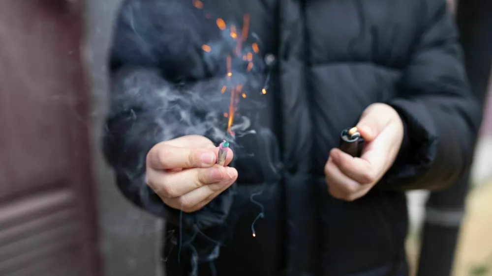 The firecracker in a hand. Boy holding a burning petard in his hand. Kid with a pyrotechnics that burns with sparks and smoke outdoors. Firecracker and lighter in hand.