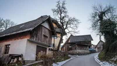 In the heart of Stara Fuzina, wooden and masonry chalets cluster along a winding lane. Textured facades, steep gabled roofs, and small windows reveal the practical alpine architecture developed for cold seasons and mountain life. Weathered boards and stone retaining walls add a sense of age and continuity, while bare trees and leftover snow signal winter. The composition highlights everyday rural character rather than tourist icons, making it ideal for illustrating Slovenian village life, vernacular building styles, heritage preservation, and the human scale of communities near Lake Bohinj. / Foto: Balkanscat Getty Images
