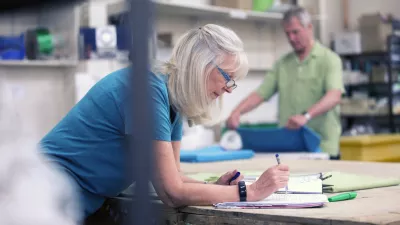 Senior businesswoman is doing paperwork in her stock warehouse. Her husband is in the background measuring fabric.