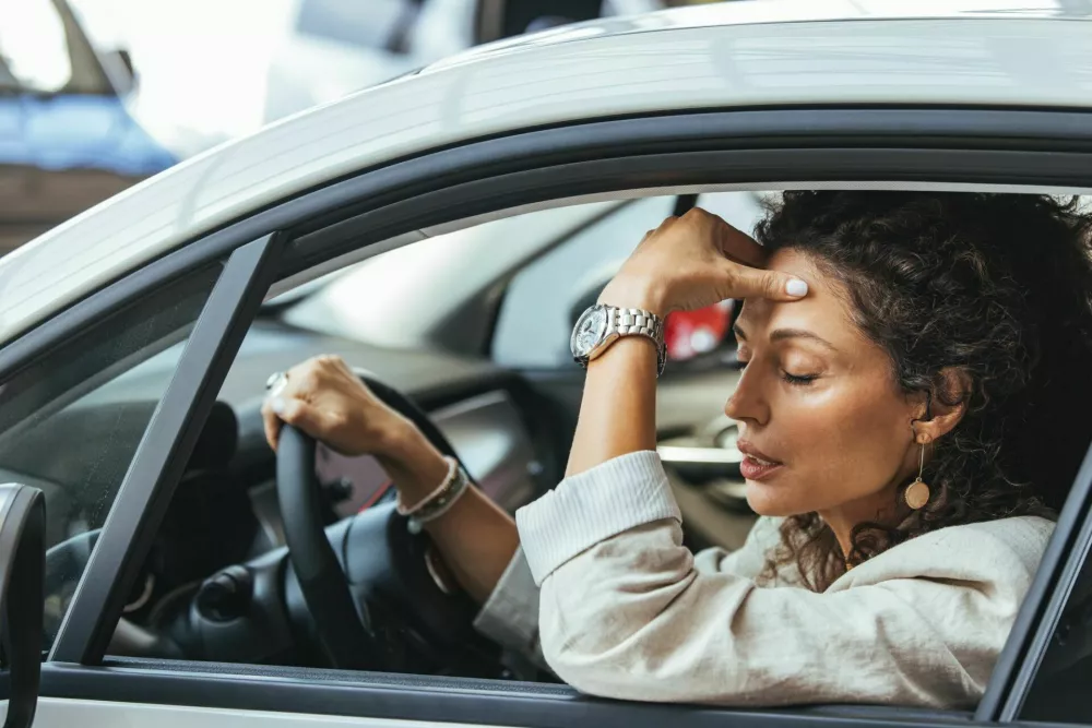 A woman sitting in her car looking stressed, caught in heavy traffic. Her hand rests on her head, showing frustration and fatigue. The urban environment adds to the tense atmosphere. / Foto: Dragana991