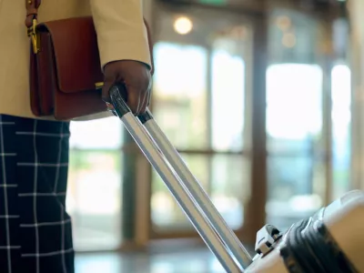 Hand of young black woman in smart casualwear with handbag pulling suitcase with luggage while walking down lounge of hotel