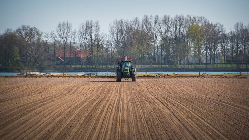 A tractor driving in the middle of arable land against trees and blue sky / Foto: Wirestock Getty Images/istockphoto