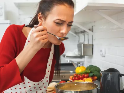 Young woman wife indoors cooking romantic dinner for husband blowing on spoon with pasta chilling out taste concentrated