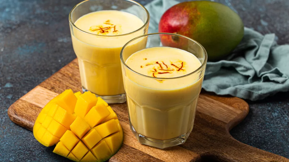 Healthy Indian Ayurveda drink mango lassi in two glasses on rustic concrete table with fresh ripe cut mango, yellow blended beverage made of mango fruit, yoghurt or milk curd and spices / Foto: Thesomegirl